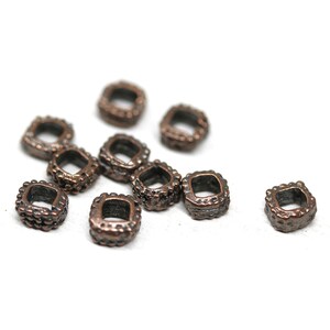 May include: A group of 10 small, square-shaped, copper-colored metal beads with a textured surface. The beads are scattered on a white background.