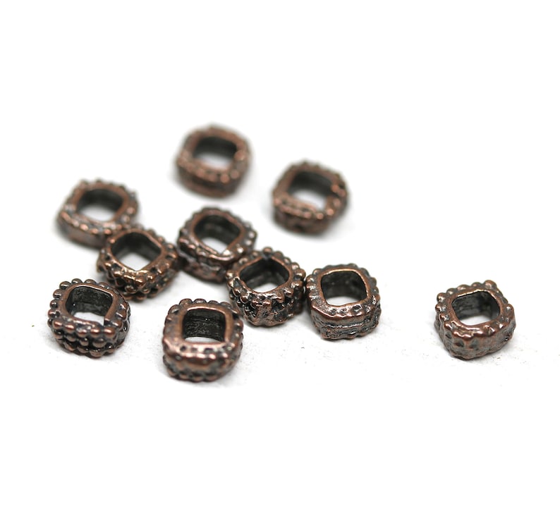 May include: A group of 10 small, square-shaped, copper-colored metal beads with a textured surface. The beads are scattered on a white background.