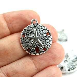 May include: Silver charm with a sand dollar design and a starfish in the center. The charm has a textured surface and four holes around the edge.