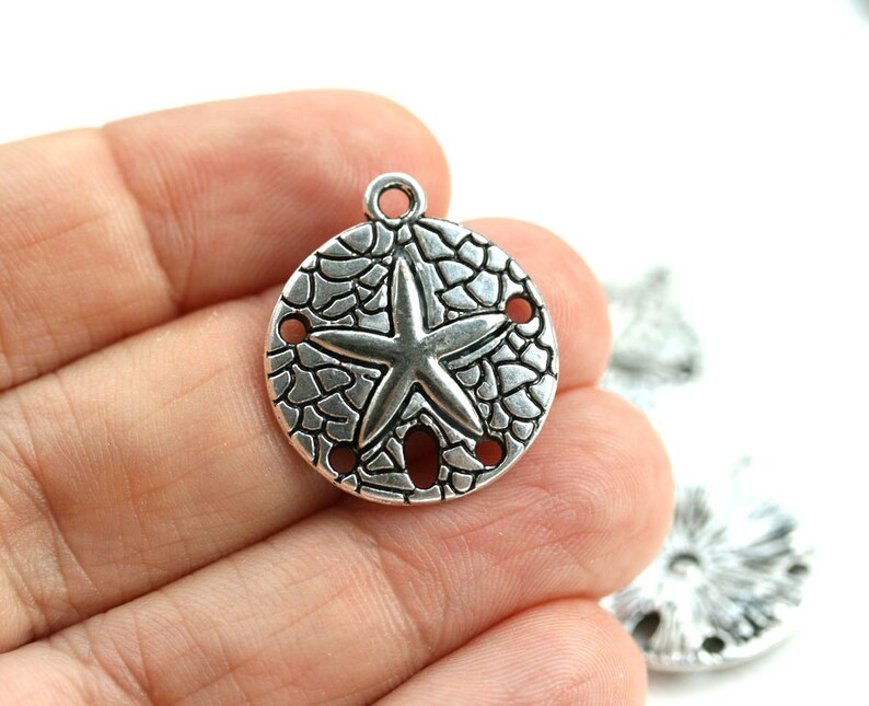 May include: Silver charm with a sand dollar design and a starfish in the center. The charm has a textured surface and four holes around the edge.