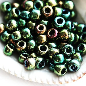 Peut inclure: Un ensemble de perles vertes irisées avec un éclat métallique, présentées dans un coquillage blanc. Les perles sont carrées avec un trou central, reflétant des teintes dorées, bleues et violettes.