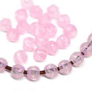 Peut inclure: Des perles roses translucides avec un motif ray&eacute; sont enfil&eacute;es sur un cordon marron. Quelques perles sont &eacute;parpill&eacute;es en arri&egrave;re-plan. Les perles ont une forme facett&eacute;e et une couleur rose clair avec des rayures blanches. Pour la cr&eacute;ation de bijoux.
