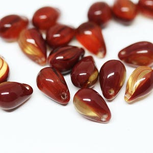 Puede incluir: Una colecci&oacute;n de cuentas de vidrio de color rojo y dorado en forma de l&aacute;grima. Las cuentas tienen una apariencia transl&uacute;cida y veteada.