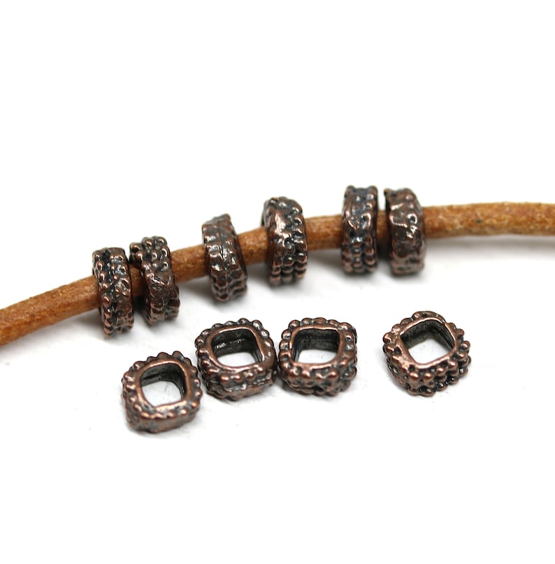 May include: A set of 10 copper-toned metal square spacer beads with a textured surface. The beads have a small hole in the center and are strung on a brown leather cord.
