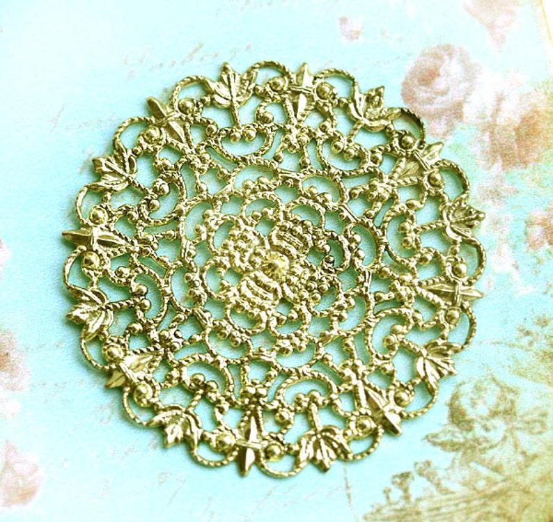May include: A gold-colored metal filigree design with a circular shape and intricate details. The design features a repeating pattern of leaves and swirls.