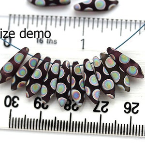 May include: A close-up of a string of dark purple glass beads with iridescent white polka dots. The beads are strung on a blue cord and are measured against a ruler, showing the size of the beads to be approximately 1/2 inch in length. The text "size demo" is visible on the ruler.