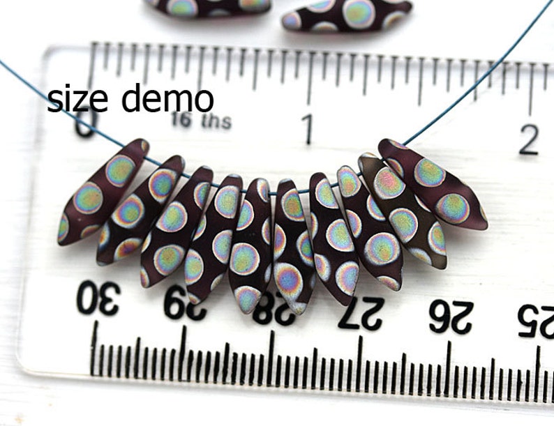 May include: A close-up of a string of dark purple glass beads with iridescent white polka dots. The beads are strung on a blue cord and are measured against a ruler, showing the size of the beads to be approximately 1/2 inch in length. The text "size demo" is visible on the ruler.