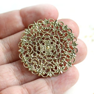 May include: A gold-toned metal filigree embellishment with a circular design. The intricate pattern features a central flower motif surrounded by delicate swirls and leaves.