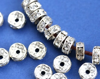May include: A close-up of silver-toned metal bead caps with clear rhinestones. The bead caps are arranged in a line and scattered on a blue surface.