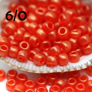 Puede incluir: Primer plano de un montón de pequeñas cuentas redondas de color rojo. Las cuentas están en una concha de mar y algunas están esparcidas sobre una superficie blanca. Las cuentas están etiquetadas como "6/0".