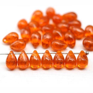 Peut inclure: Perles de verre orange en forme de goutte d'eau avec un aspect tacheté. Les perles sont enfilées sur un fil fin, avec plusieurs autres perles dispersées en arrière-plan. Ces perles conviennent à la fabrication de bijoux.