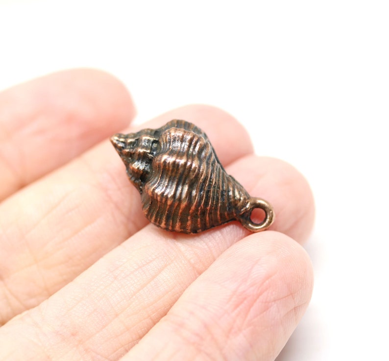 May include: A copper-coloured seashell charm with a detailed, textured surface. The charm has a small loop at the top for attaching to jewellery or other crafts.
