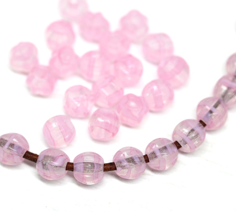 Peut inclure: Gros plan sur des perles roses translucides &agrave; rayures. Certaines sont en vrac, d'autres enfil&eacute;es sur un cordon marron. Les perles ont une forme facett&eacute;e et un motif tourbillonnant rose clair et blanc.