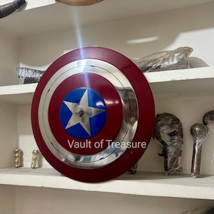 May include: A Captain America shield with a red outer ring, a silver inner ring, and a blue center with a white star. The shield is displayed on a shelf with other items.