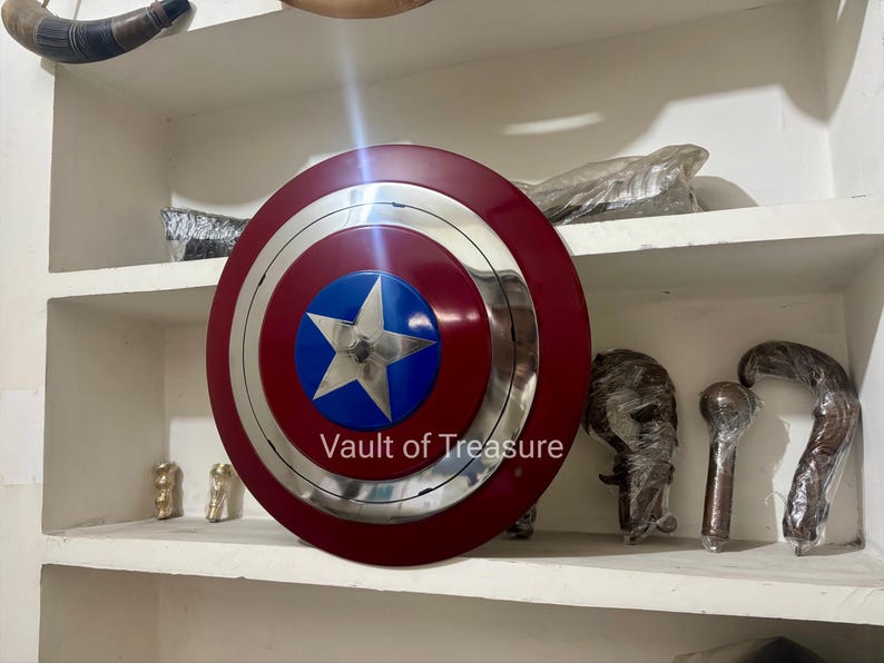 May include: A Captain America shield with a red outer ring, a silver inner ring, and a blue center with a white star. The shield is displayed on a shelf with other items.