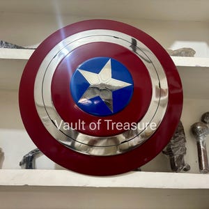 May include: A Captain America shield replica. The shield is primarily red with a silver metallic ring and a blue center featuring a white star. The text "Vault of Treasure" is visible on the shield.