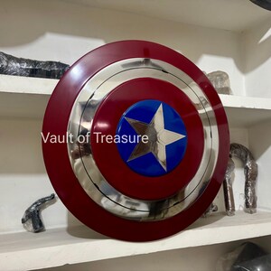 May include: A Captain America shield, featuring a red and silver concentric design with a blue circle at the center. A silver star is in the center of the blue circle. The shield is displayed on a white shelf.