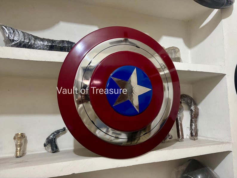 May include: A Captain America shield, featuring a red and silver concentric design with a blue circle at the center. A silver star is in the center of the blue circle. The shield is displayed on a white shelf.