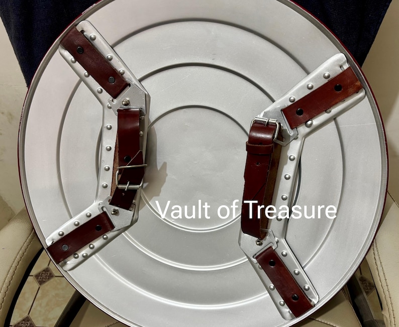 May include: A silver-colored shield with a red border and two brown leather straps. The shield has a circular design with a series of concentric circles. The text "Vault of Treasure" is visible on the shield.