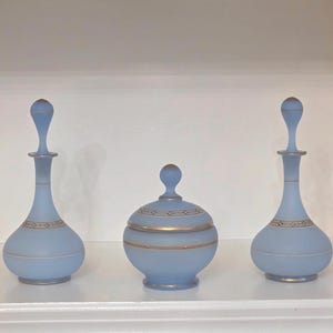 May include: Three light blue decorative glass bottles and a jar with gold accents. The bottles have a long neck and a bulbous base. The jar has a rounded shape with a lid. The set is displayed on a white shelf.