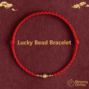 May include: A red Lucky Bead Bracelet with a gold bead in the centre. The bracelet is made of braided red cord. The text "Lucky Bead Bracelet" is displayed in gold. The background is a dark red colour.