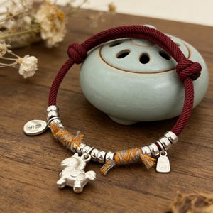 以下が含まれることがあります： シルバーのアクセントとチャームが付いた赤いコードブレスレット。ブレスレットには、小さなシルバーの馬のチャーム、シルバービーズ、小さなタグが付いています。コードは両端で結ばれています。ブレスレットは木製の表面にあります。