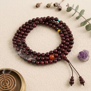 May include: A strand of dark red wooden prayer beads with a large orange bead and a tassel. A small purple crystal and incense coil are also present. The beads are likely used for meditation or spiritual practices.