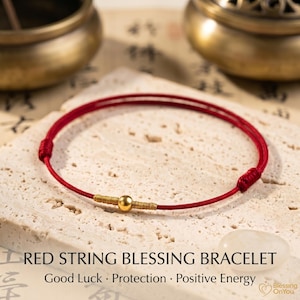 May include: A thin red string bracelet featuring a single central gold-coloured bead flanked by two small sections of gold-coloured thread wrapping. The bracelet has adjustable sliding knots on each side and the text RED STRING BLESSING BRACELET.