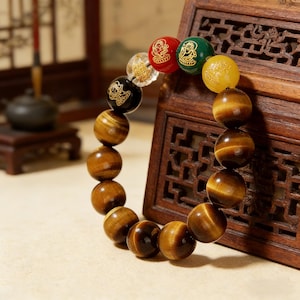 Może przedstawiać: Bransoletka z dużymi, okrągłymi kamieniami z tygrysiego oka i mniejszymi, kolorowymi koralikami akcentującymi. Koraliki akcentujące są czerwone, zielone, żółte, przezroczyste i czarne, każdy ze złotym wzorem. Bransoletka jest prezentowana na drewnianej powierzchni.