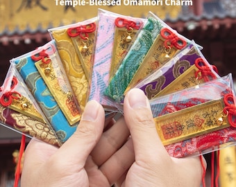 May include: A collection of Temple-Blessed Omamori charms, each in a clear plastic sleeve. The charms feature various colorful fabric designs, gold accents, and red cords. The text "Temple-Blessed Omamori Charm" is visible.