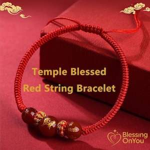 May include: A red string bracelet with three decorative beads. The beads are a deep red colour with gold accents and a gold design. The bracelet has the text "Temple Blessed Red String Bracelet". The background is red.