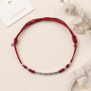 Op de afbeelding: Een rood touw armband met zilveren kralen en een cilindrisch zilveren accent. De armband is handgemaakt, zoals aangegeven door het label op de achtergrond. De armband heeft een eenvoudig ontwerp, geschikt voor dagelijks gebruik.