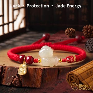 May include: A red braided bracelet with a jade dog charm, red beads, and gold accents. The bracelet also features a gold charm with Chinese characters and a fish-shaped clasp. The text "Luck Protection Jade Energy" is at the top.