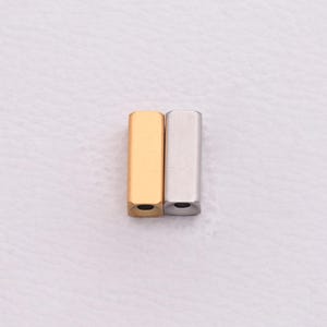 May include: Two metal bar-shaped jewelry components. One is gold-colored, the other is silver-colored. Both have a rectangular shape with rounded edges and a small hole at the bottom. The components are displayed on a white surface.