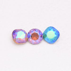 Könnte beinhalten: Drei irisierende Strasssteine in einer Reihe. Der erste ist ein abgerundetes Quadrat, der zweite ist rund und der dritte ist ein Quadrat mit abgerundeten Ecken. Jeder hat eine andere Farbe, darunter Lila, Rosa und Blau.