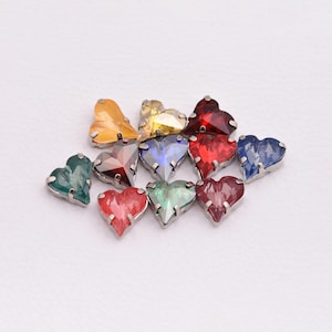 May include: A collection of heart-shaped rhinestones in various colors, including yellow, red, blue, and green. Each rhinestone has a faceted design and is set in a silver-colored metal base. The gemstones are arranged on a white surface.