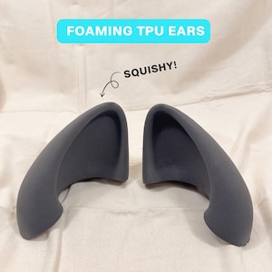 May include: A pair of dark grey, curved, foam-like TPU ears. The text FOAMING TPU EARS is displayed in a light blue box above the items, and an arrow points to one ear with the text SQUISHY! indicating the flexible material texture.