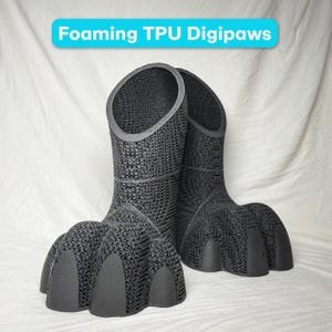 May include: A pair of black Foaming TPU Digipaws, which are tall, boot-like structures with a textured, lattice-style surface and smooth, solid toe caps. The text Foaming TPU Digipaws is displayed in a blue banner above the black paw-shaped footwear.