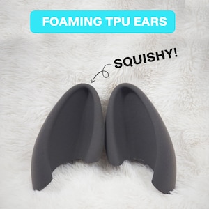 Könnte beinhalten: Ein Paar dunkelgraue Ohren aus schäumendem TPU mit einem geschwungenen, hohlen Design. Der Text FOAMING TPU EARS steht in einem hellblauen Kasten oben, und das Wort SQUISHY! mit einem Pfeil zum linken Ohr ist ebenfalls abgebildet.
