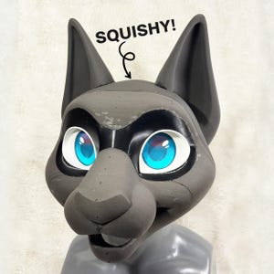 Puede incluir: Una máscara gris oscuro inspirada en un felino con grandes ojos azul brillante con bordes blancos, orejas puntiagudas y un hocico ancho. El texto SQUISHY! con una flecha hacia abajo está impreso sobre la cabeza, indicando la textura suave.