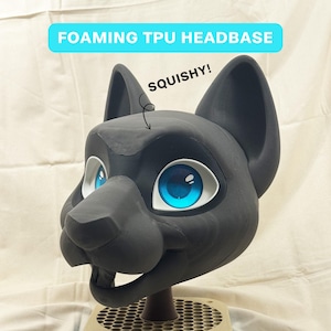 Peut inclure: Une base de tête en TPU moussant noir avec de grands yeux bleus et des accents blancs. La base de la tête a de grandes oreilles et un museau détaillé. Le texte sur l'image indique "FOAMING TPU HEADBASE" et "SQUISHY!"