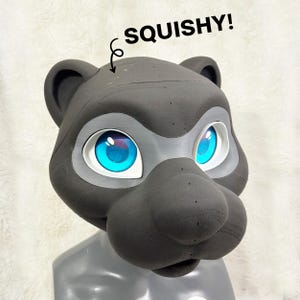 Op de afbeelding: Een donkergrijze berenhoofd met grote, helderblauwe ogen en een witte en grijze oogomtrek. Het woord "SQUISHY!" staat boven het hoofd. De berenhoofd maakt waarschijnlijk deel uit van een kostuum of cosplay-artikel.