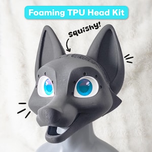 Pode incluir: Um kit de cabeça de TPU espumoso cinza-escuro com uma máscara de raposa com olhos grandes azul-brilhantes e bordas brancas. O texto "Foaming TPU Head Kit" aparece no topo e a palavra "squishy!" está escrita com uma seta para a orelha.