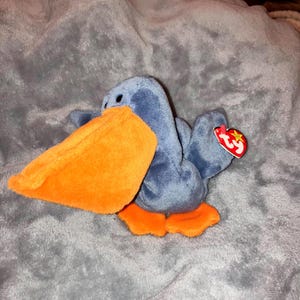 Könnte beinhalten: Ein Stofftier-Pelikan mit blauem Körper, orangefarbenem Schnabel und orangefarbenen Füßen. Ein rotes Etikett ist angebracht. Der Pelikan liegt auf einem grauen, strukturierten Untergrund.