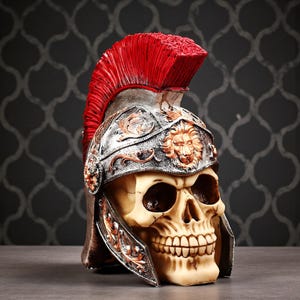 Könnte beinhalten: Ein dekorativer Schädel mit einem römisch anmutenden Helm mit rotem Kamm. Der Helm ist silberfarben mit Bronze- und Goldakzenten und weist ein Löwenemblem auf. Der Schädel hat ein detailliertes, realistisches Aussehen.