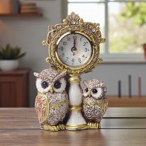 Puede incluir: Un reloj decorativo con temática de búhos, con un marco dorado y una esfera blanca. Dos figuras de búhos sostienen el reloj, con detalles intrincados en marrón, beige y dorado. El reloj marca aproximadamente la 1:00.