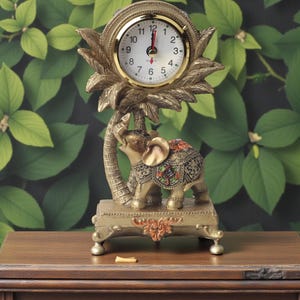 May include: A gold-coloured decorative clock with an elephant statue base. The clock face is white with black numerals and gold trim. The elephant is decorated with colourful patterns and stands on a rectangular base. The clock measures approximately 15 cm in height.