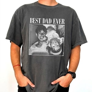 Op de afbeelding: Donkergrijze t-shirt met een zwart-wit foto-opdruk. De foto toont een man en een kind, met de tekst "BEST DAD EVER" boven de afbeelding. Het shirt heeft korte mouwen en een ronde hals.