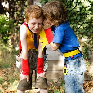May include: Two children dressed as superheroes, one in a red and yellow costume with a silver cape and the other in a blue shirt with a yellow belt and a red and yellow arm band. The child in the blue shirt is whispering in the ear of the child in the red and yellow costume.