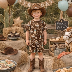 May include: A young child in a brown cowboy hat, shirt, and shorts stands in front of a birthday party setup. The outfit has a repeating pattern. Hay bales, balloons, and a banner that says "Happy Birthday" are in the background. A sign reads "Howdy Partner!"